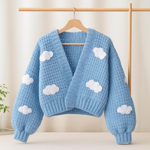 May include: A light blue, open-front cardigan with white cloud appliqués. The sweater has a textured knit pattern, long, puffy sleeves, and ribbed cuffs. The cardigan is displayed on a wooden hanger.