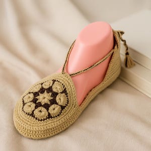 Puede incluir: Zapatilla de ganchillo beige con un diseño floral marrón oscuro en la puntera. La zapatilla tiene una correa en el tobillo y un pequeño detalle de borla. La zapatilla se muestra en un pie rosa. Talla 37-38.