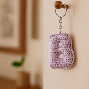 Puede incluir: Un llavero de ganchillo con la letra "B" en color lavanda, colgado en una pared. La letra está hecha de hilo y tiene dos agujeros. El llavero está sujeto a un anillo y una cadena de metal plateado.