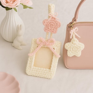 Op de afbeelding: Een crèmekleurig, gehaakt fotolijstje met een roze strik en een bijpassende stervormige bedel. Een roze handtas met een stervormige bedel hangt in de buurt. Een witte vaas met roze rozen en een wit konijnenbeeldje staan op de achtergrond.