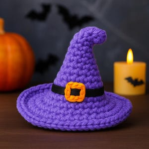 Pode incluir: Um chapéu de bruxa roxo de crochê com uma faixa preta e fivela laranja. O chapéu está sobre uma superfície de madeira com uma vela acesa e uma abóbora no fundo. Decoração com tema de Halloween.