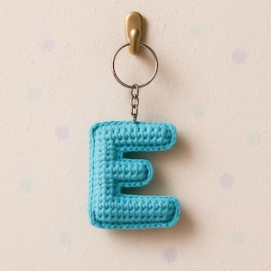 Puede incluir: Un llavero con la letra "E" de ganchillo, de color azul turquesa, colgado de un gancho dorado. La letra está hecha de un hilo grueso y tiene un diseño redondeado y cuadrado. El llavero está unido a un anillo y una cadena de metal.