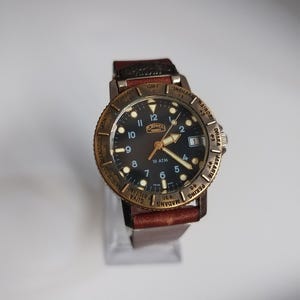 K&ouml;nnte beinhalten: Eine Armbanduhr mit schwarzem Zifferblatt, goldfarbener L&uuml;nette und braunem Lederarmband. Das Zifferblatt der Uhr hat wei&szlig;e Ziffern und Zeiger, mit dem Markennamen "CAMEL". Auf der L&uuml;nette sind St&auml;dtenamen eingraviert.