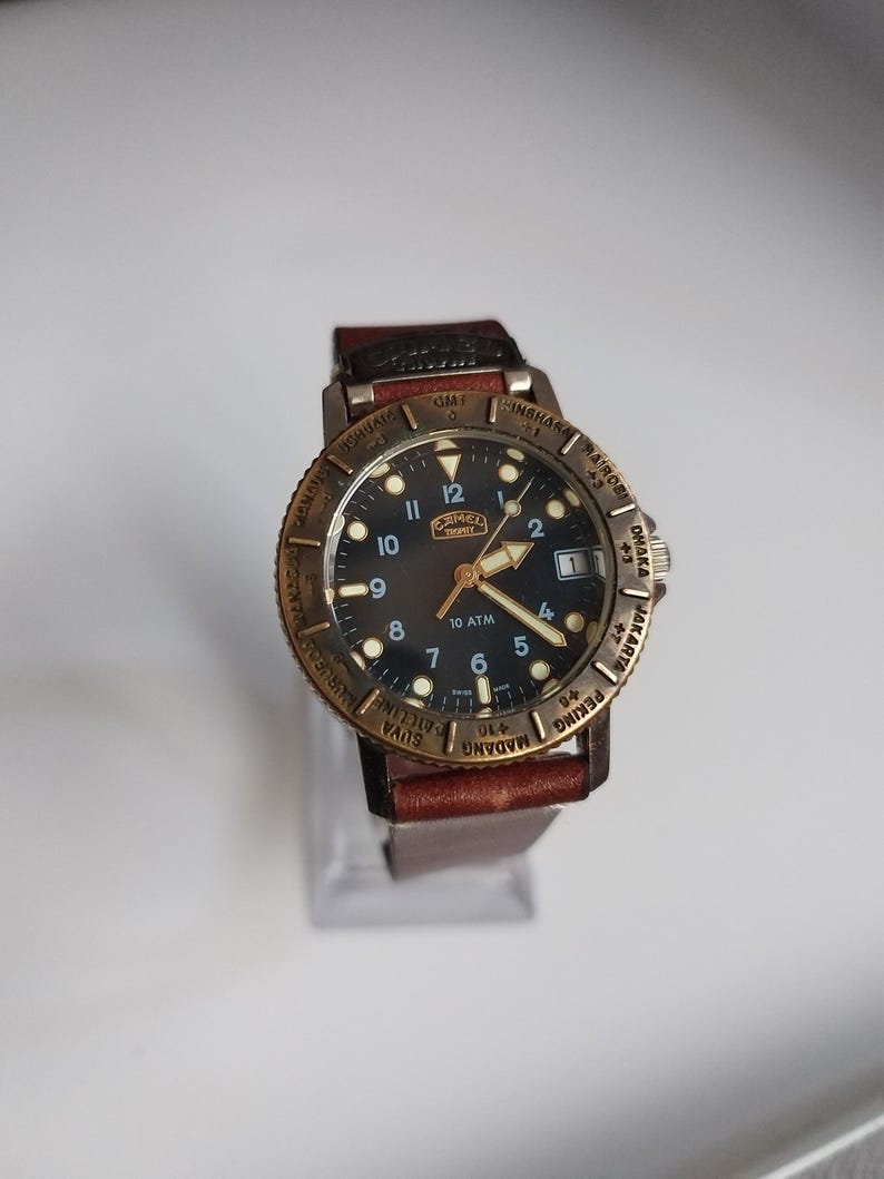 K&ouml;nnte beinhalten: Eine Armbanduhr mit schwarzem Zifferblatt, goldfarbener L&uuml;nette und braunem Lederarmband. Das Zifferblatt der Uhr hat wei&szlig;e Ziffern und Zeiger, mit dem Markennamen "CAMEL". Auf der L&uuml;nette sind St&auml;dtenamen eingraviert.