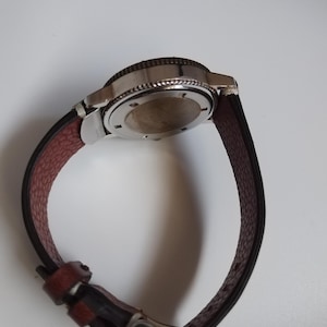 K&ouml;nnte beinhalten: Eine Armbanduhr mit einem Edelstahlgeh&auml;use und einem braunen Lederarmband. Die Uhr hat ein dunkles Zifferblatt und eine silberne Schlie&szlig;e. Das Armband ist etwa 23 cm lang.