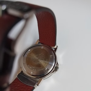 K&ouml;nnte beinhalten: Eine silberfarbene Armbanduhr mit einem braunen Lederarmband. Die R&uuml;ckseite der Uhr ist sichtbar und zeigt Text und Markierungen. Das Armband ist strukturiert, tiefbraun und das Uhrengeh&auml;use ist silberfarben. Ein klassisches Accessoire.