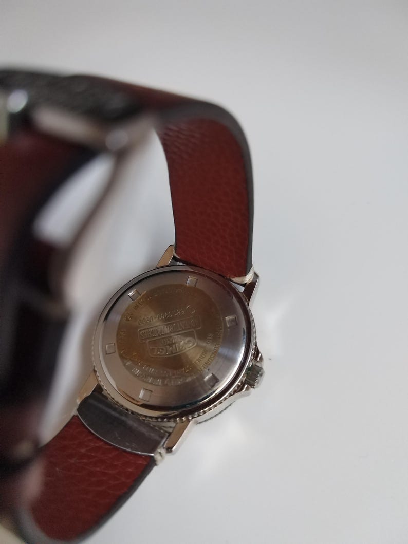 K&ouml;nnte beinhalten: Eine silberfarbene Armbanduhr mit einem braunen Lederarmband. Die R&uuml;ckseite der Uhr ist sichtbar und zeigt Text und Markierungen. Das Armband ist strukturiert, tiefbraun und das Uhrengeh&auml;use ist silberfarben. Ein klassisches Accessoire.