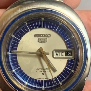 Può includere: Primo piano di un orologio da polso automatico Seiko 5 vintage. L'orologio ha una cassa color argento, un quadrante color crema con dettagli blu e un display giorno-data. Le lancette e gli indici dell'orologio sono dorati. L'orologio è contrassegnato "Automatic 21 Jewels".