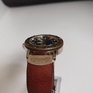 K&ouml;nnte beinhalten: Eine Armbanduhr mit schwarzem Zifferblatt, goldfarbener L&uuml;nette und braunem Lederarmband. Die Uhr hat ein klassisches Design mit sichtbaren Zeigern und Markierungen. Das Armband ist mit dem Wort "CAMEL" versehen.