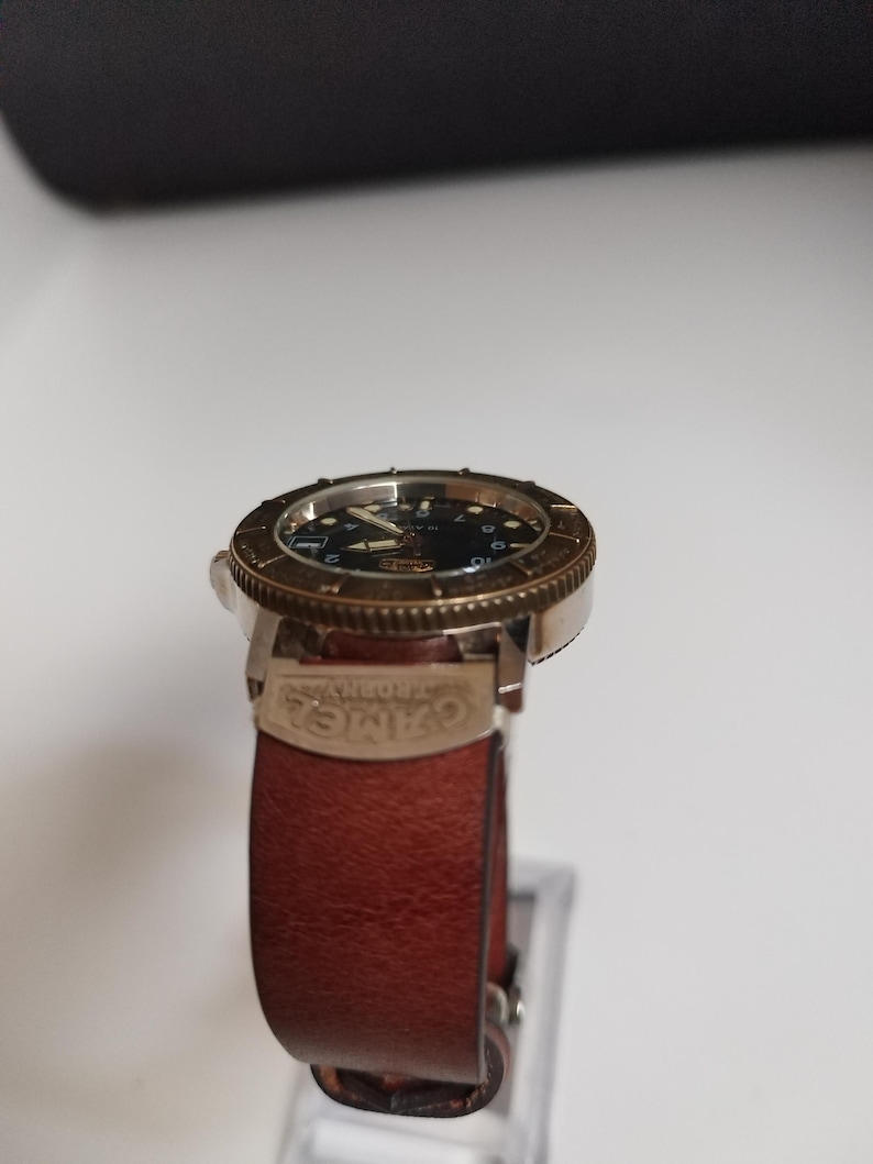 K&ouml;nnte beinhalten: Eine Armbanduhr mit schwarzem Zifferblatt, goldfarbener L&uuml;nette und braunem Lederarmband. Die Uhr hat ein klassisches Design mit sichtbaren Zeigern und Markierungen. Das Armband ist mit dem Wort "CAMEL" versehen.