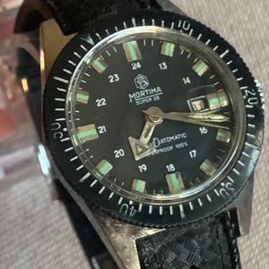 May include: A vintage Mortima Super 28 wristwatch with a black dial and bezel. The watch features a black leather strap and a date display at the 3 o'clock position. The dial has luminous markers and hands.