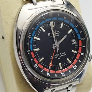 Puede incluir: Un reloj de pulsera automático Seiko Navigator Timer de acero inoxidable con esfera negra, ventana de fecha en la posición de las tres y bisel giratorio con escala de 24 horas en azul y rojo. El reloj incluye una correa de eslabones metálicos.
