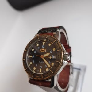 K&ouml;nnte beinhalten: Eine Vintage-Armbanduhr mit braunem Zifferblatt, goldfarbener L&uuml;nette und braunem Lederarmband. Das Zifferblatt der Uhr zeigt arabische Ziffern und den Markennamen "CAPEL". Die Uhr wird auf einem transparenten St&auml;nder pr&auml;sentiert.