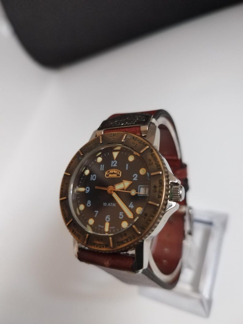 K&ouml;nnte beinhalten: Eine Vintage-Armbanduhr mit braunem Zifferblatt, goldfarbener L&uuml;nette und braunem Lederarmband. Das Zifferblatt der Uhr zeigt arabische Ziffern und den Markennamen "CAPEL". Die Uhr wird auf einem transparenten St&auml;nder pr&auml;sentiert.