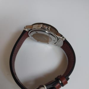K&ouml;nnte beinhalten: Eine Edelstahl-Armbanduhr mit einem schwarzen und braunen Lederarmband. Die Uhr hat eine silberne L&uuml;nette und ein dunkles Zifferblatt. Das Armband ist etwa 23 cm lang. Die Uhr ist f&uuml;r den t&auml;glichen Gebrauch konzipiert.