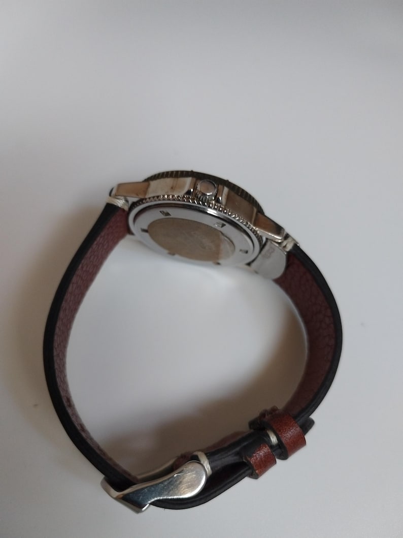 K&ouml;nnte beinhalten: Eine Edelstahl-Armbanduhr mit einem schwarzen und braunen Lederarmband. Die Uhr hat eine silberne L&uuml;nette und ein dunkles Zifferblatt. Das Armband ist etwa 23 cm lang. Die Uhr ist f&uuml;r den t&auml;glichen Gebrauch konzipiert.