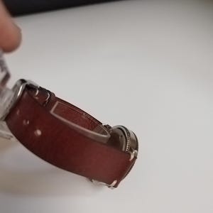 K&ouml;nnte beinhalten: Eine weinrote Uhr mit einem silberfarbenen Metallarmband. Das Zifferblatt der Uhr ist rund und hat eine strukturierte Oberfl&auml;che. Das Armband besteht aus einem glatten, festen Material.