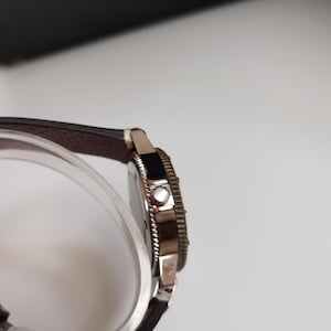 K&ouml;nnte beinhalten: Eine silberfarbene Armbanduhr mit einem dunkelbraunen Lederarmband. Das Zifferblatt der Uhr ist schwarz und die L&uuml;nette hat ein strukturiertes Design. Die Uhr wird auf einem transparenten, gebogenen St&auml;nder pr&auml;sentiert.