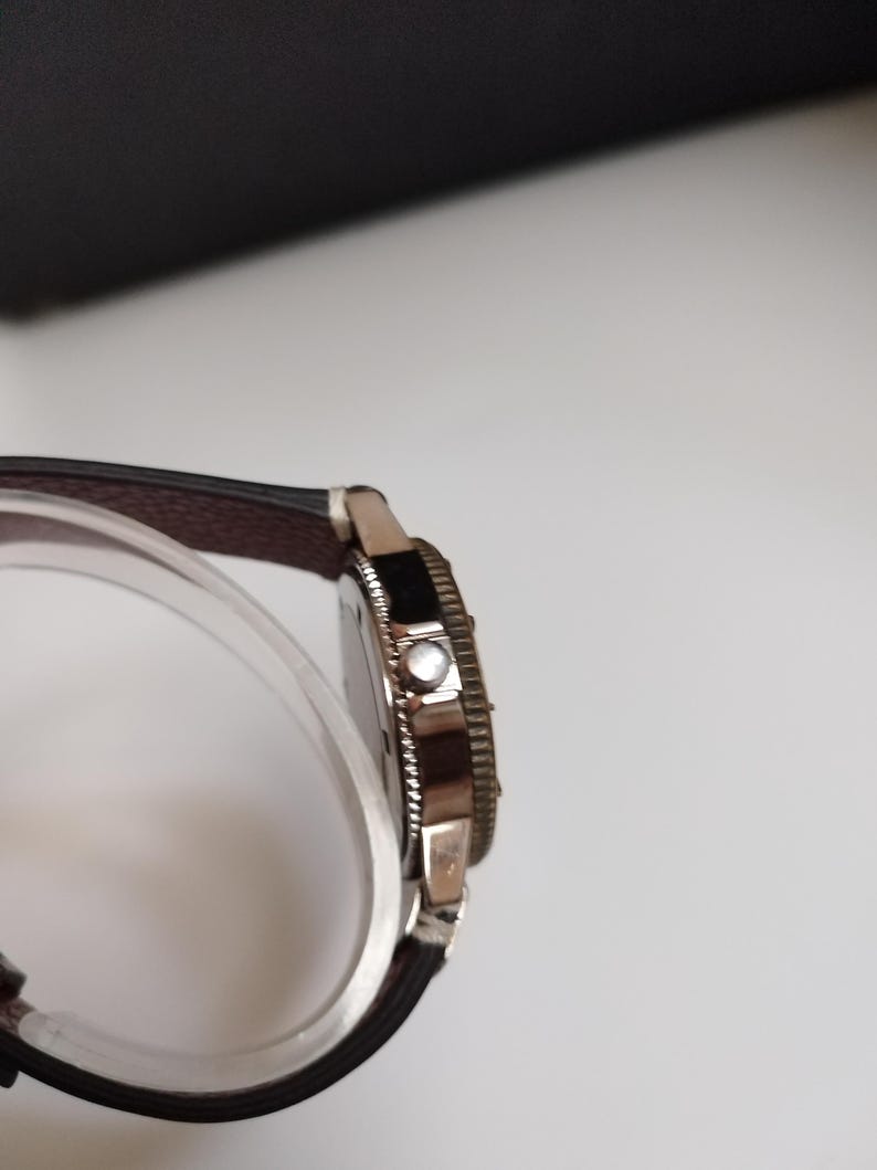K&ouml;nnte beinhalten: Eine silberfarbene Armbanduhr mit einem dunkelbraunen Lederarmband. Das Zifferblatt der Uhr ist schwarz und die L&uuml;nette hat ein strukturiertes Design. Die Uhr wird auf einem transparenten, gebogenen St&auml;nder pr&auml;sentiert.