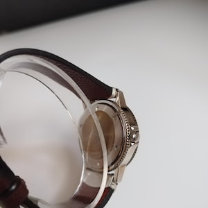 K&ouml;nnte beinhalten: Eine Edelstahl-Armbanduhr mit einem dunkelbraunen Lederarmband. Die Uhr hat eine silberne L&uuml;nette und einen transparenten Kunststoff-Armbandhalter. Das Zifferblatt der Uhr ist nicht sichtbar.