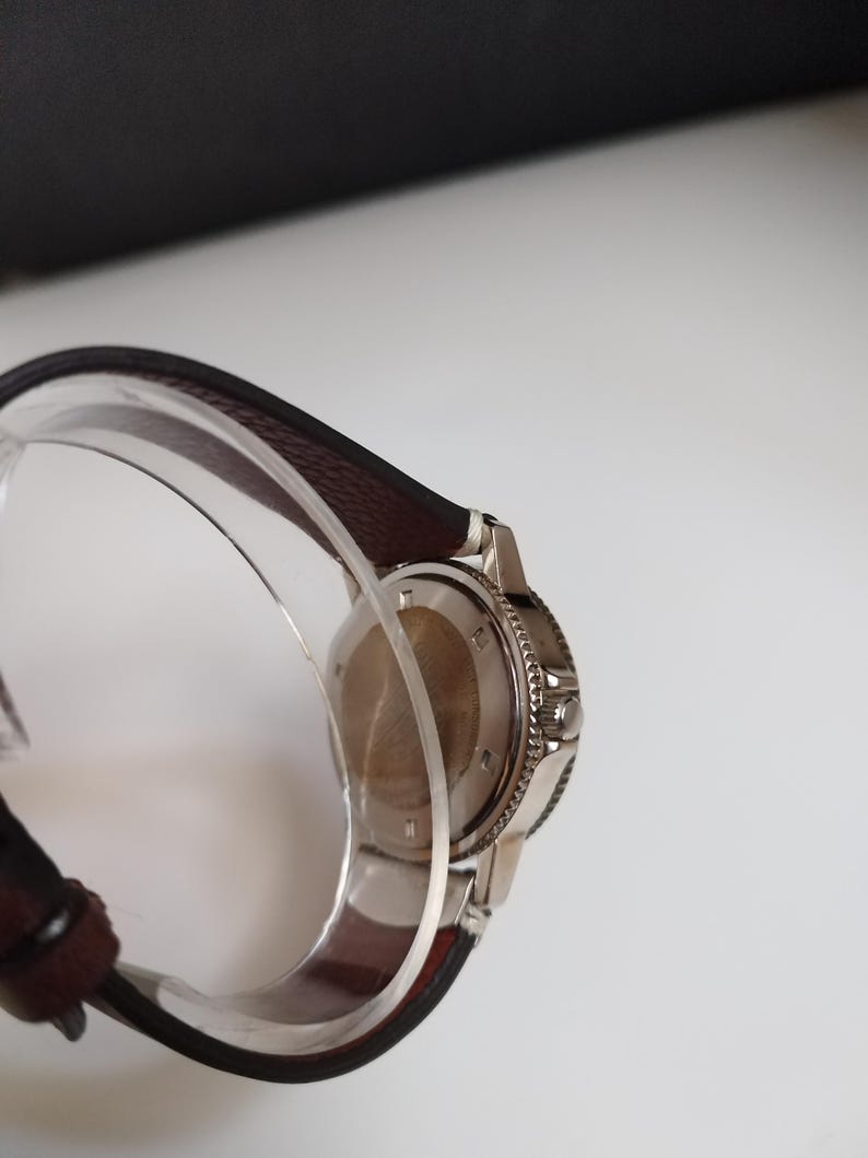 K&ouml;nnte beinhalten: Eine Edelstahl-Armbanduhr mit einem dunkelbraunen Lederarmband. Die Uhr hat eine silberne L&uuml;nette und einen transparenten Kunststoff-Armbandhalter. Das Zifferblatt der Uhr ist nicht sichtbar.
