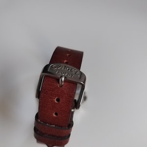 K&ouml;nnte beinhalten: Nahaufnahme eines braunen Leder-Uhrenarmbands mit silberner Schnalle. Das Armband hat ein klassisches Design mit sichtbaren N&auml;hten und einem Metallverschluss. Die Schnalle ist mit dem Text "CAMEL TROPHY" graviert.