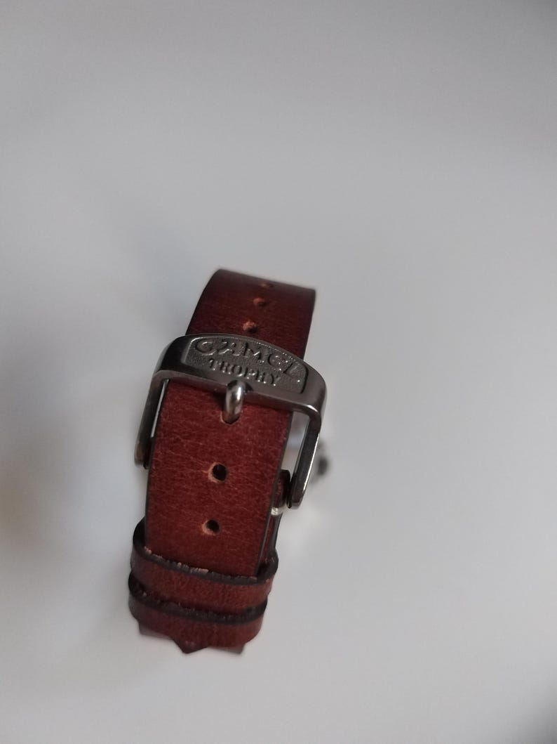 K&ouml;nnte beinhalten: Nahaufnahme eines braunen Leder-Uhrenarmbands mit silberner Schnalle. Das Armband hat ein klassisches Design mit sichtbaren N&auml;hten und einem Metallverschluss. Die Schnalle ist mit dem Text "CAMEL TROPHY" graviert.