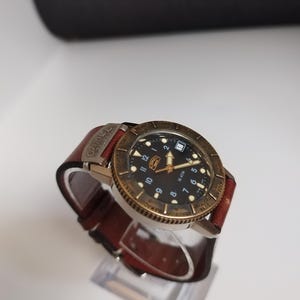 K&ouml;nnte beinhalten: Eine Armbanduhr mit schwarzem Zifferblatt, goldfarbener L&uuml;nette und braunem Lederarmband. Die Uhr hat wei&szlig;e Ziffern und Zeiger mit Datumsanzeige bei 3 Uhr. Das Armband wird mit einer Schnalle befestigt. Die Uhr wird auf einem transparenten St&auml;nder pr&auml;sentiert.