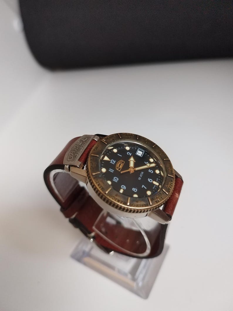 K&ouml;nnte beinhalten: Eine Armbanduhr mit schwarzem Zifferblatt, goldfarbener L&uuml;nette und braunem Lederarmband. Die Uhr hat wei&szlig;e Ziffern und Zeiger mit Datumsanzeige bei 3 Uhr. Das Armband wird mit einer Schnalle befestigt. Die Uhr wird auf einem transparenten St&auml;nder pr&auml;sentiert.
