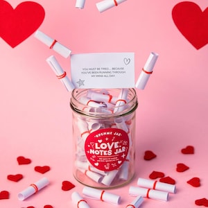 Może przedstawiać: Szklany słoik z etykietą "Love Notes Jar" wypełniony zwiniętymi notatkami, czerwonymi sercami i pływającymi notatkami. Słoik ma czerwoną etykietę z białym tekstem. Tło jest różowe.