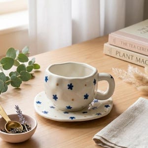 May include: A white ceramic teacup and saucer set with blue flower and gold dot designs. The cup has a rounded shape and a thick handle. The saucer is round with a matching pattern. A small bowl with a gold spoon and lavender is in the foreground.