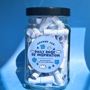 Puede incluir: Un tarro de cristal transparente lleno de rollos de papel, asegurados con bandas azules. El tarro tiene una tapa negra y una etiqueta azul que dice "OPENME JAR. DAILY DOSE OF INSPIRATION" con gráficos florales y de nubes.