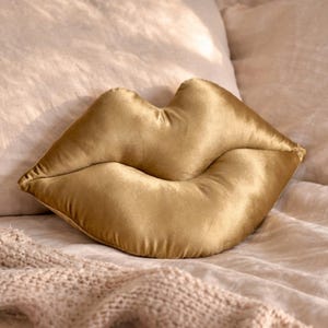 May include: A gold velvet lip-shaped cushion. The cushion is placed on a bed with neutral-toned bedding. The cushion's shape is a stylized representation of a pair of lips. The bed also has a knitted throw.