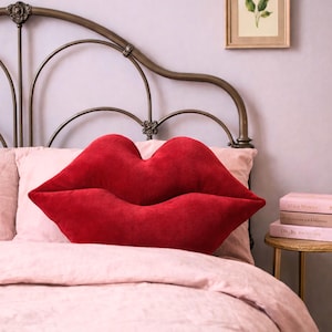 May include: A vibrant red lip-shaped cushion sits on a bed with pink bedding. The cushion has a soft, velvety texture. A decorative metal headboard and a small side table with books are also visible in the image.