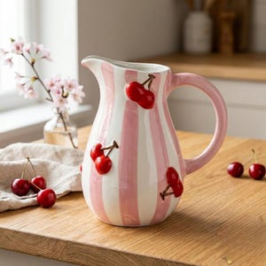 Puede incluir: Jarra de cerámica a rayas rosas y blancas, con diseños de cerezas rojas en relieve. La jarra tiene un asa curva y está sobre una superficie de madera. Aproximadamente 20 cm de altura.