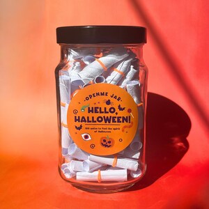 May include: A clear glass jar with a black lid filled with rolled-up white notes secured with orange rubber bands. A round orange label reads "OPENME JAR. HELLO, HALLOWEEN!" with Halloween-themed graphics. The jar is set against an orange background.