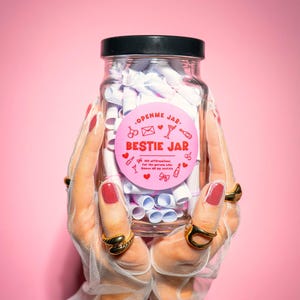 May include: A clear glass jar with a black lid labeled "BESTIE JAR" in pink, filled with rolled-up white paper. The jar is held by hands wearing gold rings and white gloves, set against a pink backdrop. The label includes the text "100 affirmations for the person who knows all my secrets."