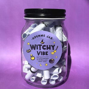 May include: A clear glass jar with a black lid filled with rolled-up white paper scrolls. A round sticker on the jar reads "Witchy Vibe" with illustrations of a witch's hat, broom, and cauldron. The jar contains 50 affirmations to awaken your inner witch.