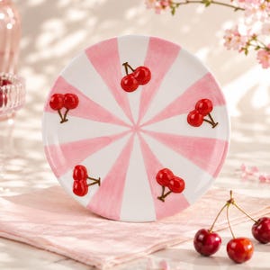 Puede incluir: Un plato redondo de cerámica con un patrón de rayas radiales en blanco y rosa claro. El plato está decorado con cuatro conjuntos de motivos de cerezas rojas brillantes en relieve, colocados a intervalos alrededor del centro.