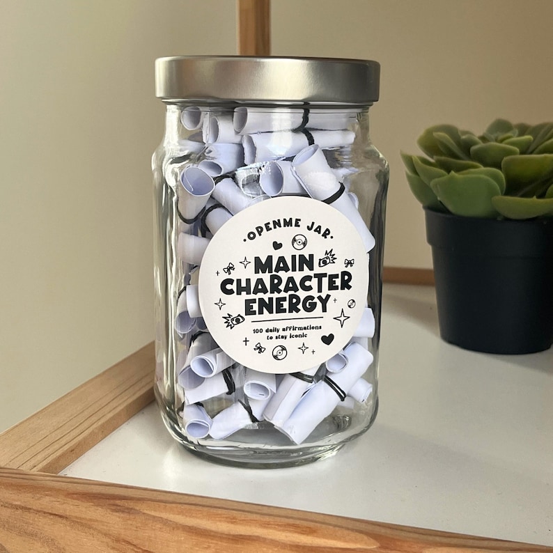 May include: A clear glass jar with a silver lid filled with rolled-up white paper scrolls. A white label on the jar reads "OPENME JAR. MAIN CHARACTER ENERGY" with additional text and decorative elements. A small potted plant is visible.