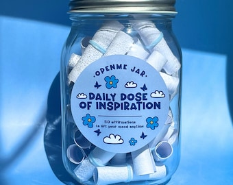 May include: A clear glass jar with a silver lid filled with rolled-up white paper scrolls. A blue sticker on the jar reads ".Open Me Jar. Daily Dose of Inspiration" with floral and cloud graphics. The jar contains 50 affirmations.