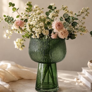 May include: A green glass vase with a ribbed design, holding a bouquet of flowers. The flowers include pink carnations and white blossoms with green foliage. The vase has a wide, rounded top and a pedestal base.
