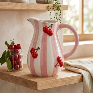 Pode incluir: Jarro de cerâmica com listras rosa e brancas, adornado com cerejas vermelhas. O jarro tem uma alça curva e um bico. Um frasco de cerejas está em uma prateleira de madeira. É um item decorativo de cozinha.