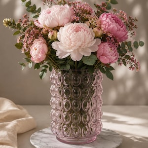 Puede incluir: Un ramo de peonías rosas y otras flores en un jarrón de cristal rosa. El jarrón tiene un patrón texturizado de círculos. Las flores son de varios tonos de rosa y tienen hojas verdes. El jarrón está sobre una superficie similar al mármol.