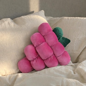 Puede incluir: Un cojín en forma de uva rosa con un acento de hoja verde. El cojín está hecho de un material suave y aterciopelado y está diseñado para parecerse a un racimo de uvas. Descansa sobre una superficie de lino blanco.