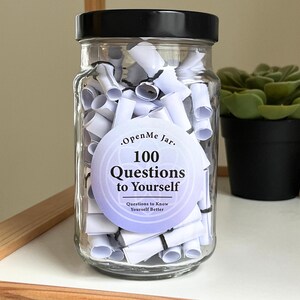 May include: A clear glass jar with a black lid filled with rolled-up white paper. A circular label on the jar reads "Open Me Jar. 100 Questions to Yourself. Questions to Know Yourself Better."