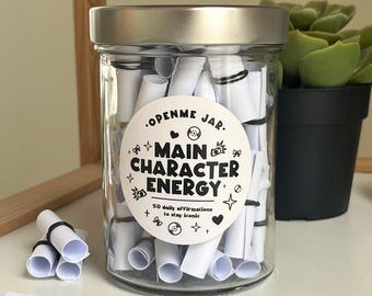 May include: A clear glass jar with a silver lid filled with rolled-up white paper scrolls. A round sticker on the jar reads "OPENME JAR. MAIN CHARACTER ENERGY" with additional text and graphics. Several scrolls are outside the jar. A potted plant is in the background.