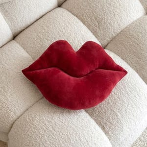 May include: A red, lips-shaped cushion made from a soft, velvety fabric. The cushion is a deep shade of red and is designed in the shape of a pair of lips. A decorative item for a bed or sofa.