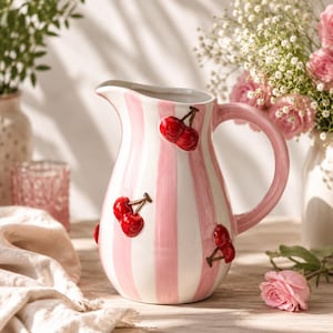 Peut inclure: Un pichet en céramique orné de rayures verticales blanches et rose clair. Le pichet est décoré de motifs de cerises rouges en relief avec des tiges brunes réparties sur toute la surface extérieure et comprend une anse incurvée.