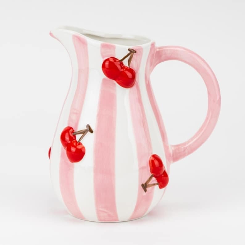 May include: A ceramic pitcher with a pink and white striped pattern. The pitcher features a curved handle and is adorned with red cherry accents. The pitcher is designed for serving beverages and adds a decorative touch to any table setting.